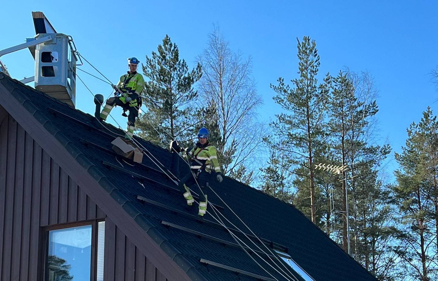 Kaksi valjastettua asentajaa asentamassa aurinkopaneeleita katolle nostokurki näkyvissä